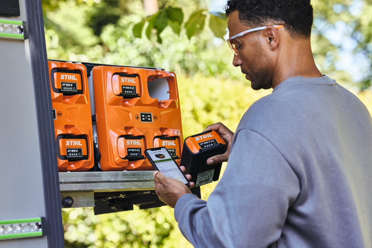 In the foreground is a man who has opened the STIHL connected app. In the background is the STIHL multiple charger AL 301-4