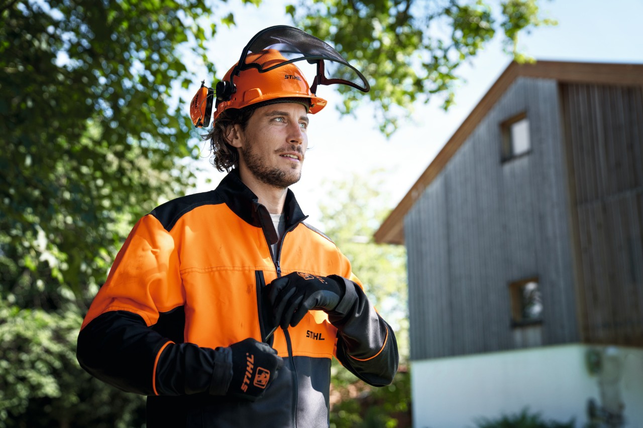 Man with orange and black STIHL protective equipment including helmet and gloves looks confidently out of the picture, in the background a house and trees 